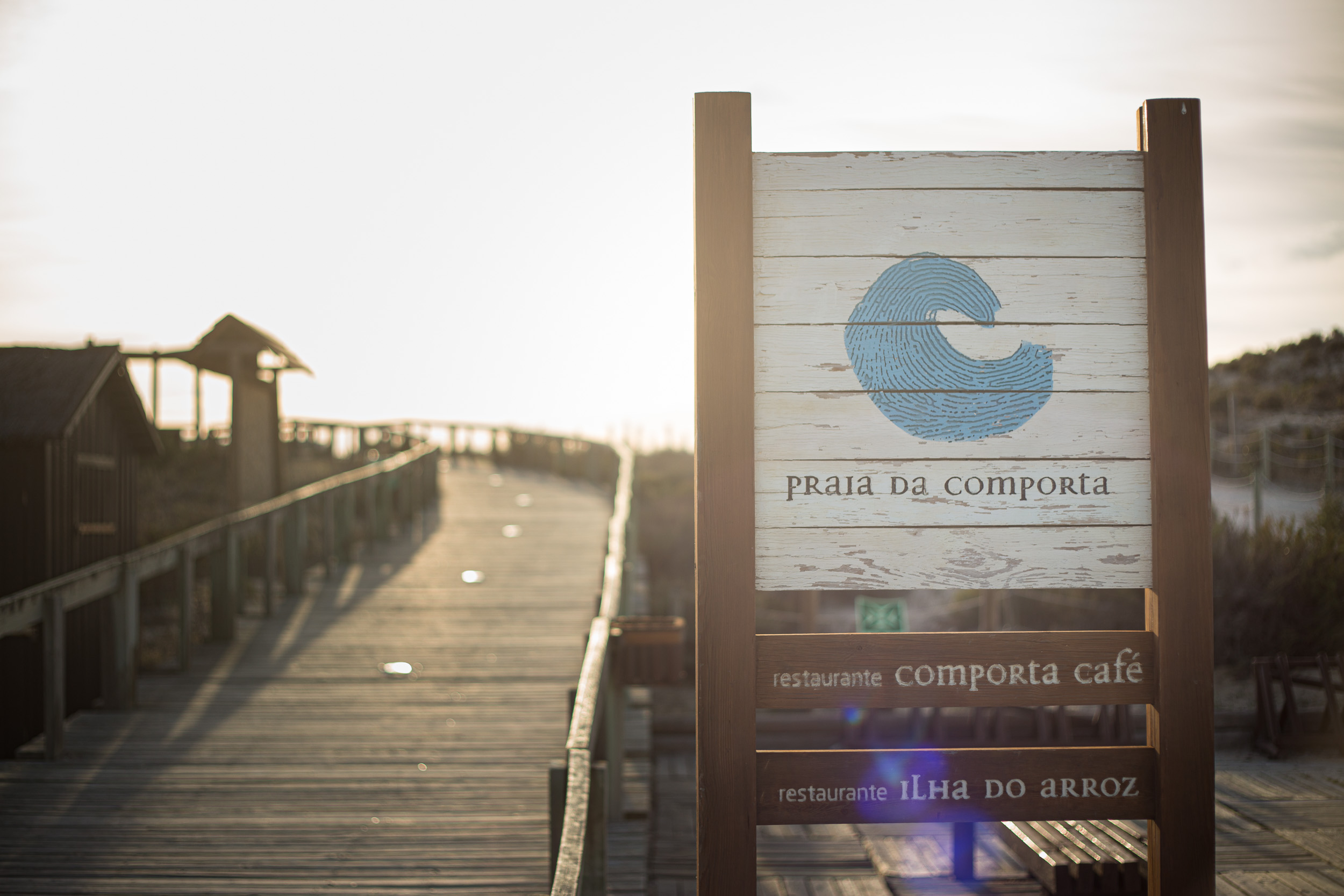 Un panneau en bois sur le ponton menant à Praia da Comporta avec des directions pour Comporta Café et Ilha do Arroz en contre-jour doré.