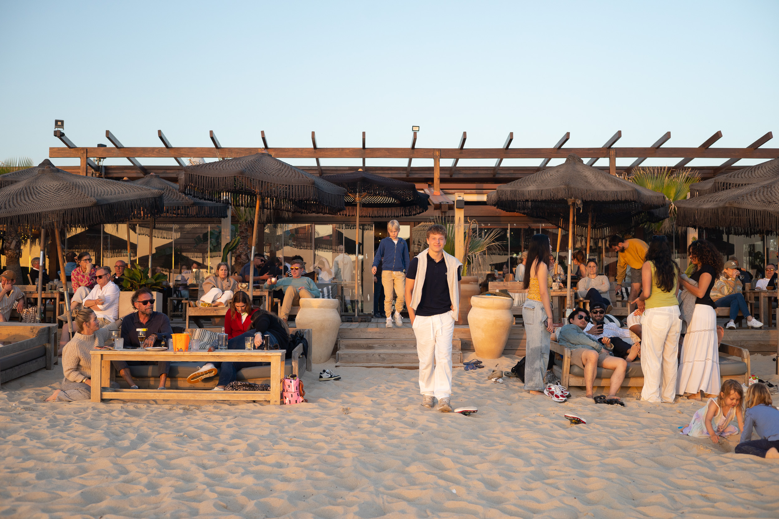 Gäste entspannen auf der Sandterrasse des Comporta Café unter dunklen Sonnenschirmen im späten Nachmittagslicht.