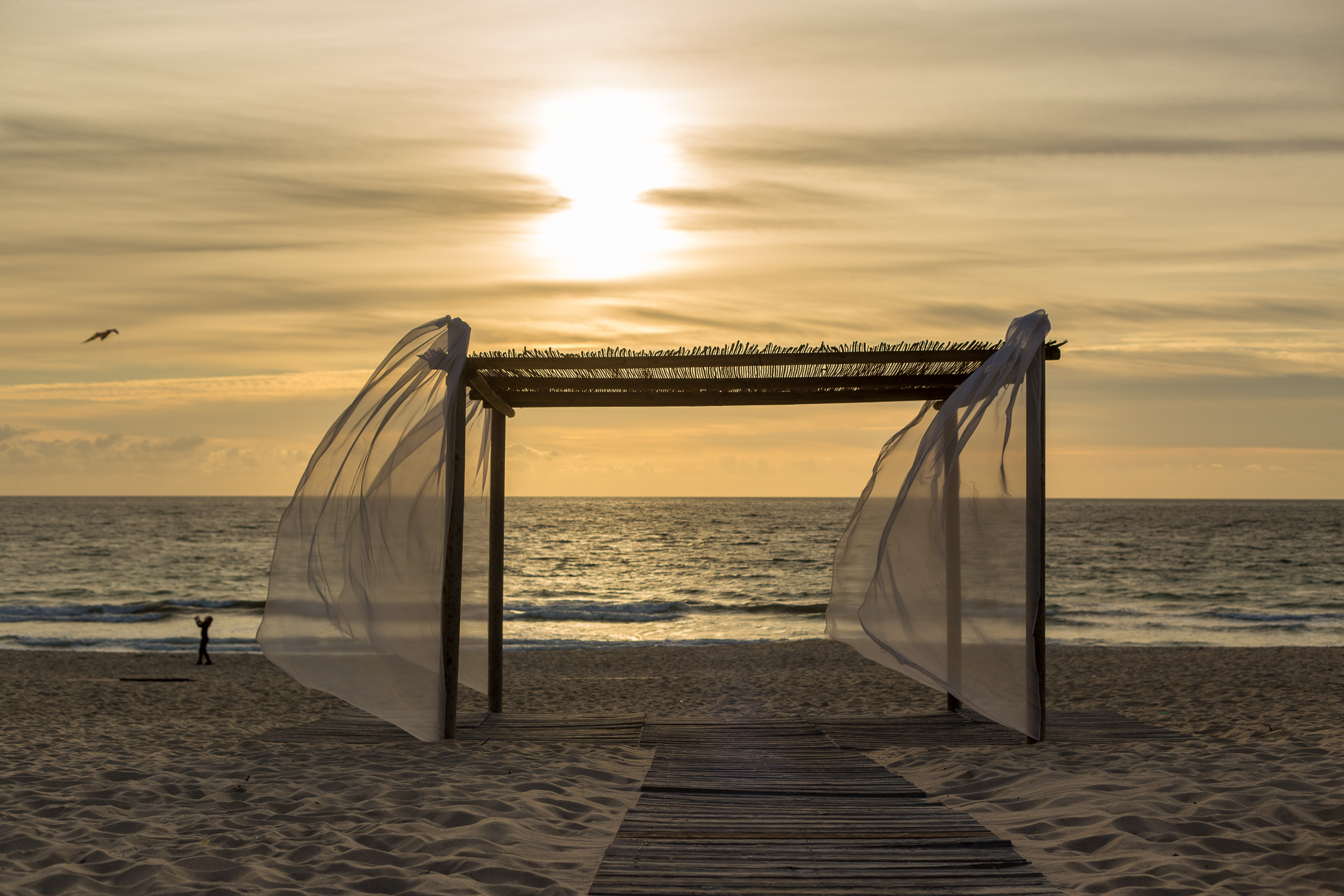 Une pergola en bois avec des tissus blancs flottants sur la plage dans la lumière dorée du coucher de soleil sur la mer.