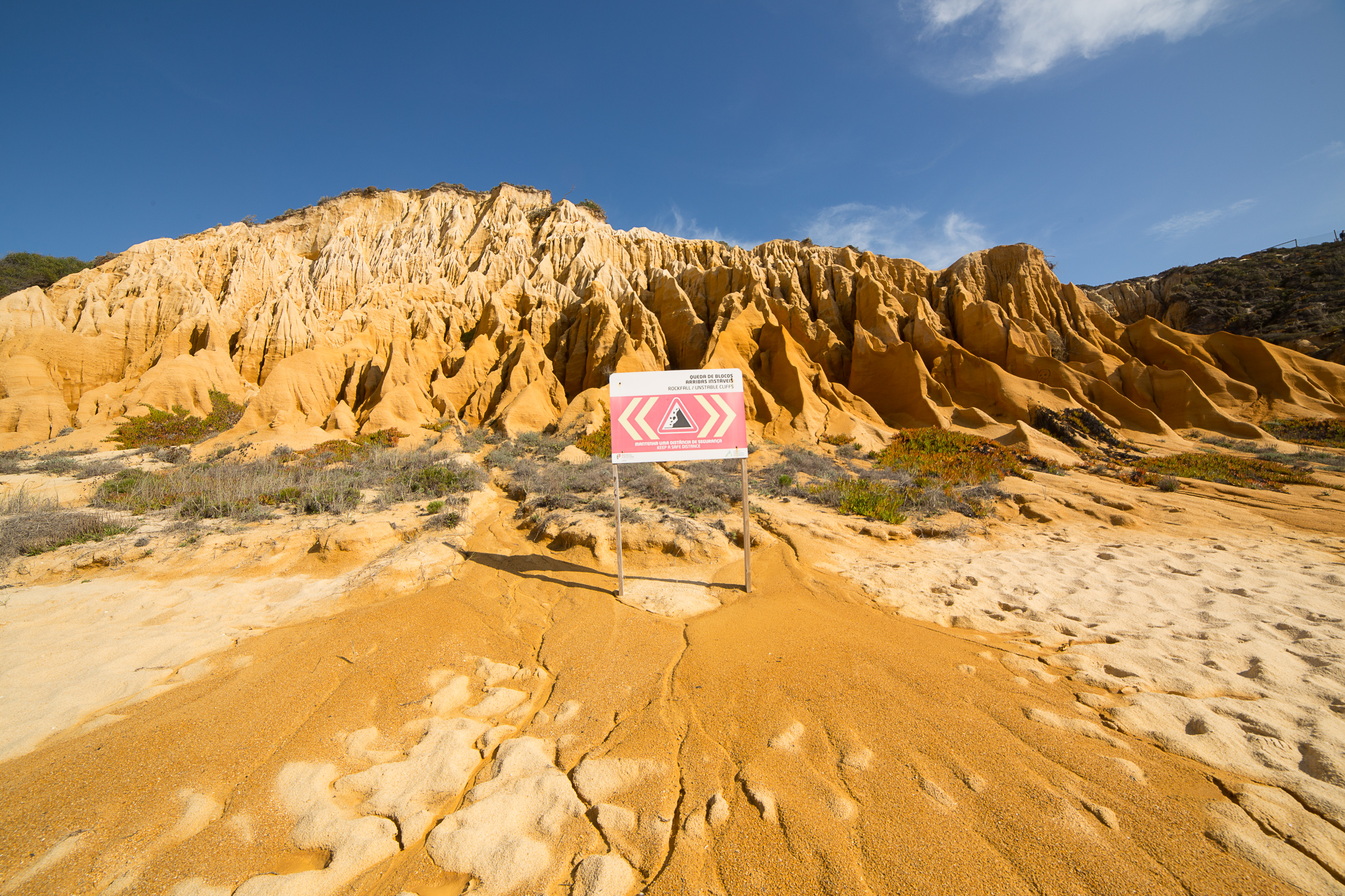 Des falaises stratifiées dans des tons orange et ocre rejoignent l'eau, au premier plan se trouve un panneau d'avertissement rouge sur la plage.